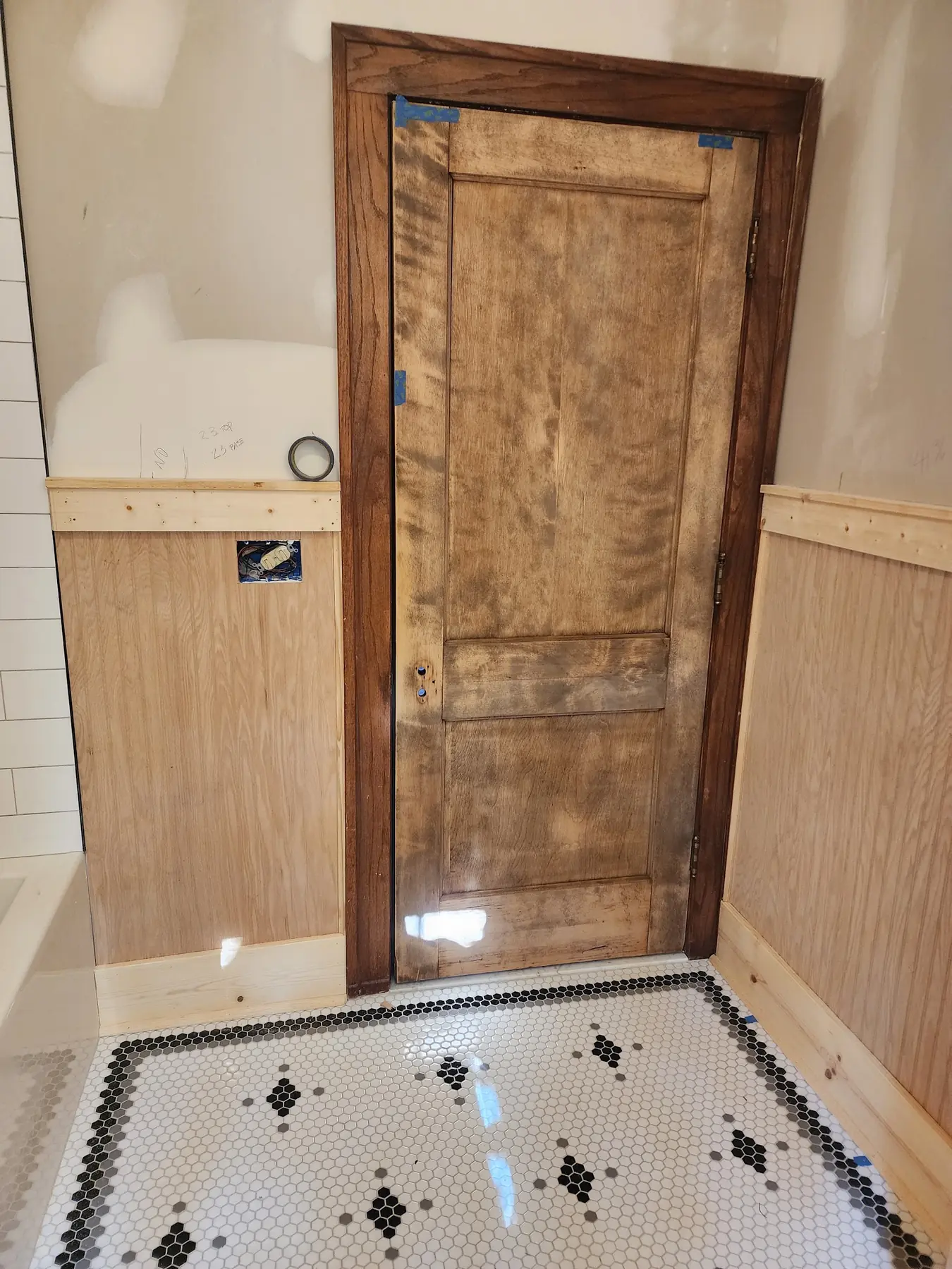 Bathroom door mid-renovation: blotchy bare wood in a new frame, unpainted beadboard and a hex tile floor